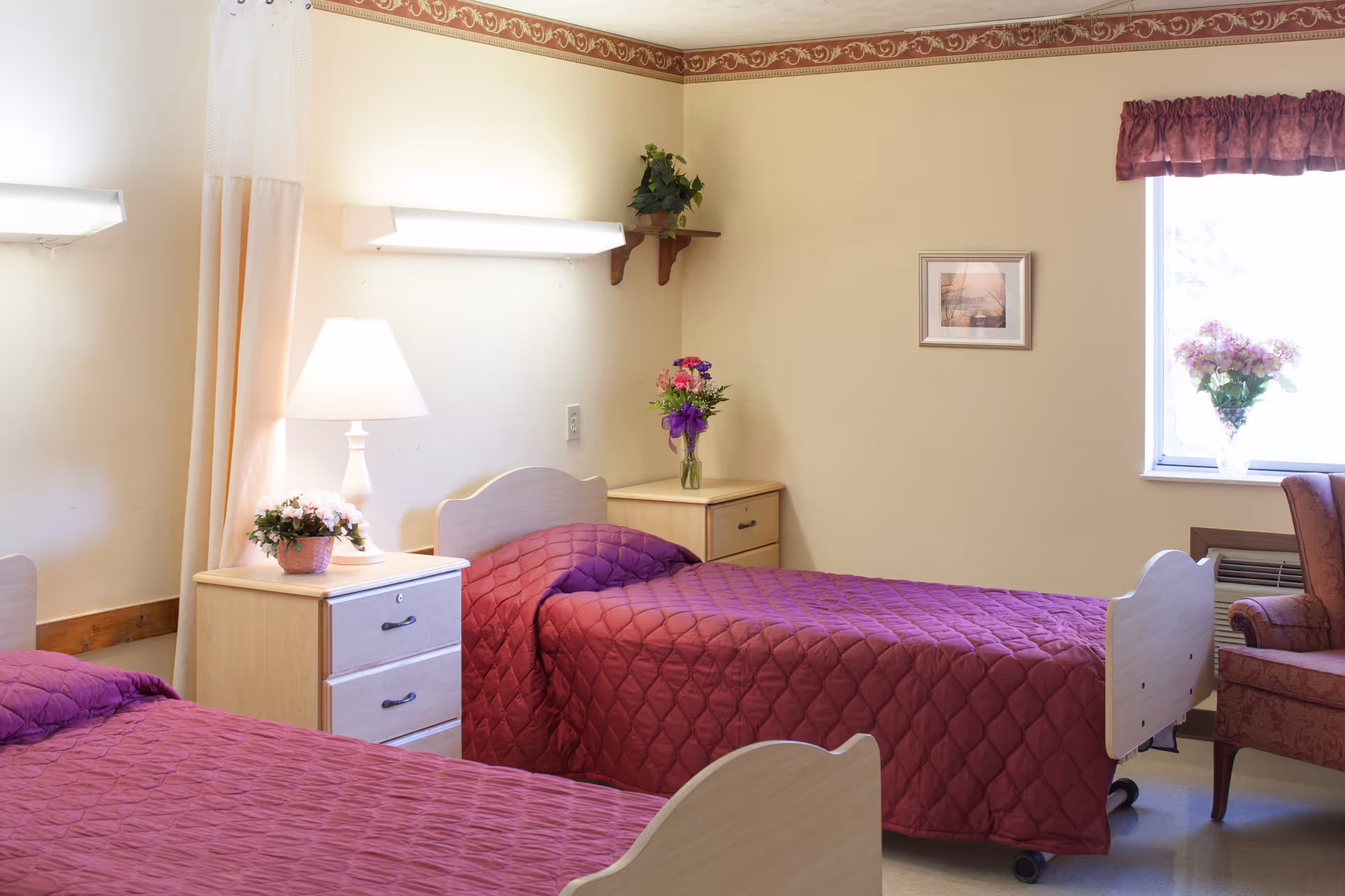 A bright senior-care bedroom with two twin beds covered in burgundy quilted bedspreads, nightstands with lamps and flowers, a window, and a chair.