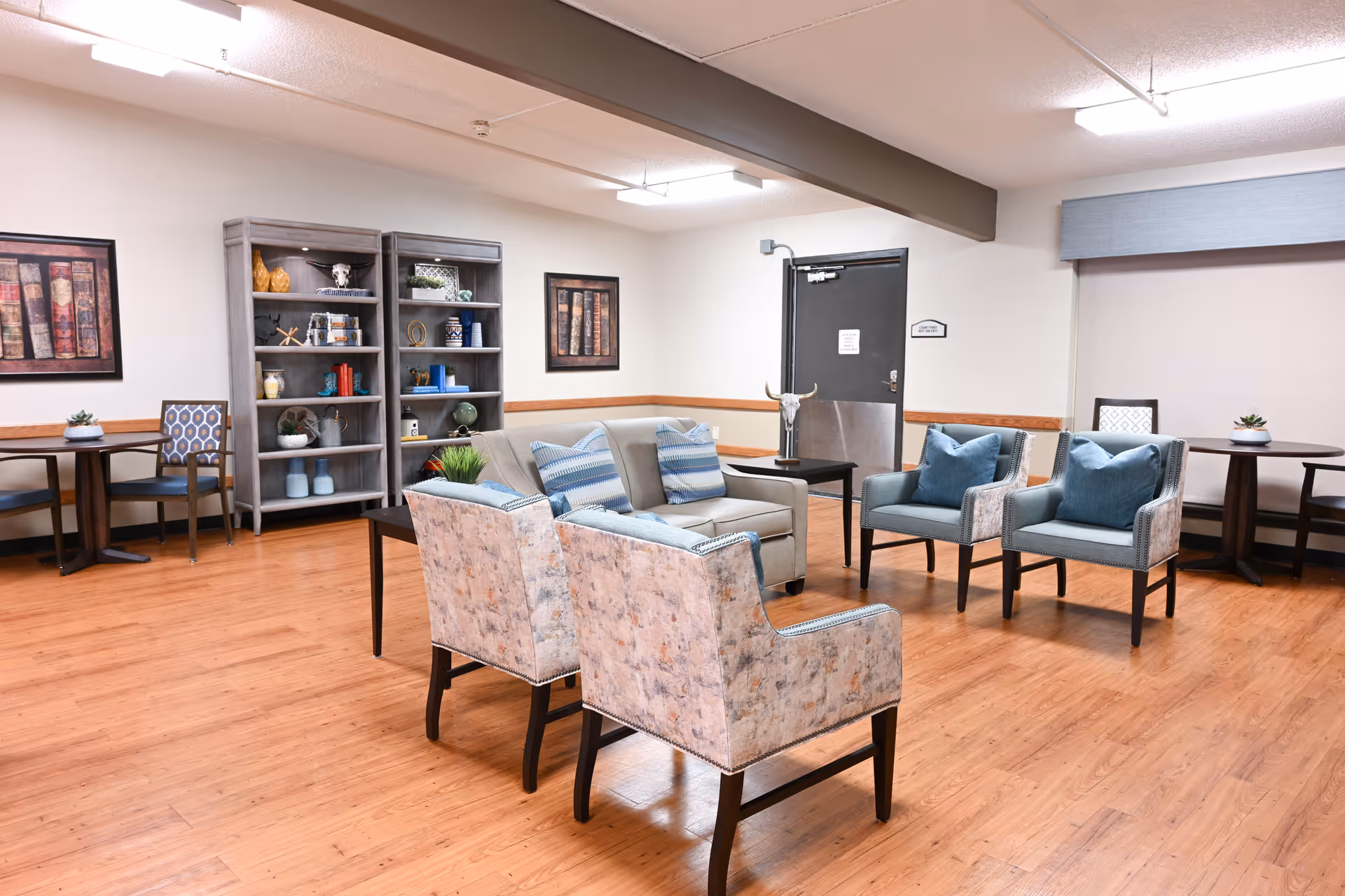 A well-lit common area with wooden flooring featuring a seating arrangement of a sofa with blue striped cushions, two patterned armchairs, and two blue cushioned chairs. There are two small round tables with chairs along the walls, decorative shelves with various ornaments, and framed artwork on the walls. A black door is visible in the background.