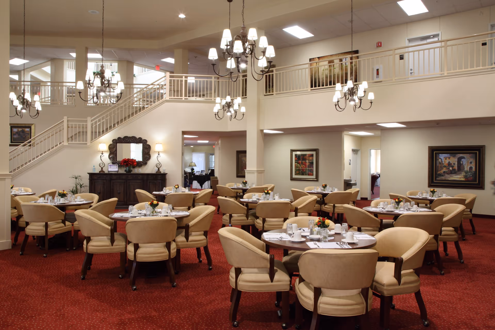 A spacious dining room with multiple round tables set with white tableware and small floral centerpieces. The room features beige upholstered chairs, red carpet flooring, cream-colored walls, and several chandeliers hanging from the ceiling. There is a staircase with a railing leading to an upper level, and framed artwork on the walls.
