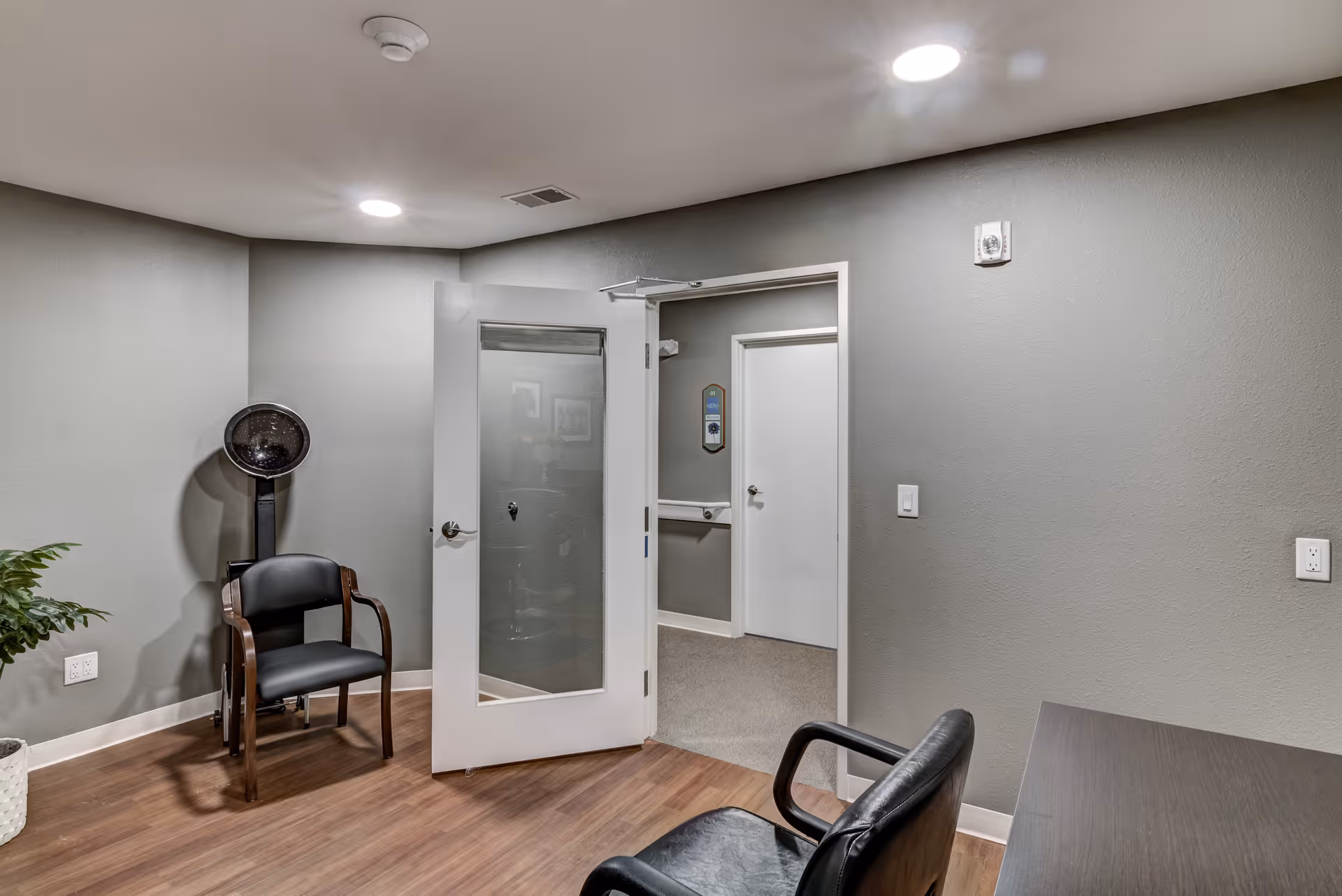 Interior room with gray walls and wood flooring featuring two black chairs, one with wooden arms and the other with leather upholstery, a hair dryer hood mounted on a stand, a white door with a glass panel partially open leading to a hallway with carpeted floor and another closed white door.