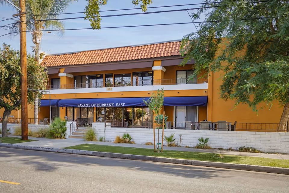 Front exterior of the Savant of Burbank East building with a blue awning over the entrance, balconies on the second floor, and a tiled roof.