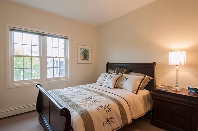 A cozy bedroom with a wooden bed frame and a neatly made bed featuring a beige and green floral patterned comforter and matching pillows. There is a wooden nightstand with a lamp, books, and an alarm clock. A window with white blinds lets in natural light, and a framed picture hangs on the wall.