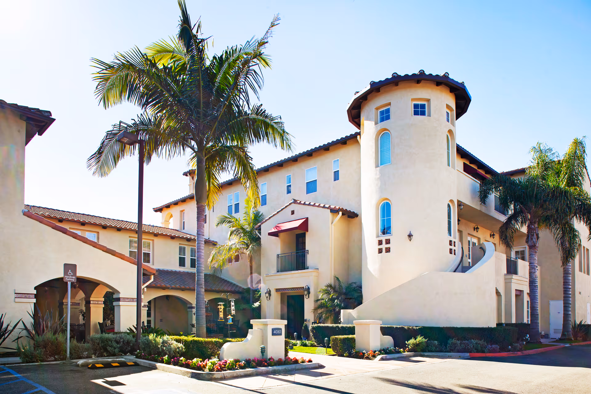 Exterior view of a multi-story senior living facility with beige stucco walls, red tile roofing, palm trees, and a well-maintained garden area in front. The building features a rounded tower-like structure and balconies with railings.
