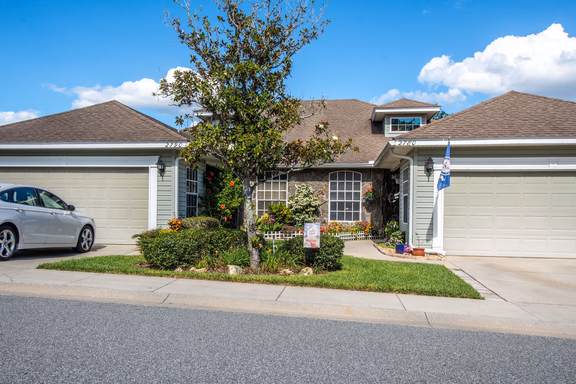 Front exterior of a single-story townhouse unit with two garages, a central entrance, small landscaped yard and a parked silver car.