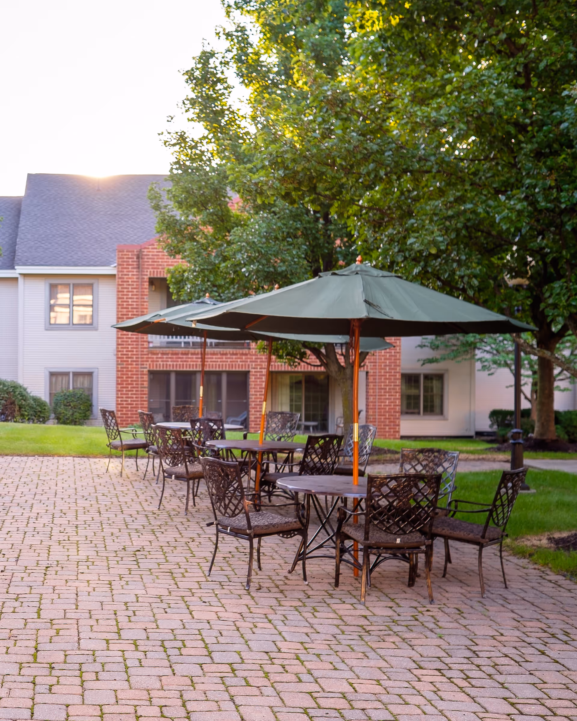 Outdoor patio area with multiple round tables and metal chairs arranged on a brick-paved surface. Each table has a large green umbrella providing shade. The background shows a building with red brick and beige siding, along with green trees and grass.