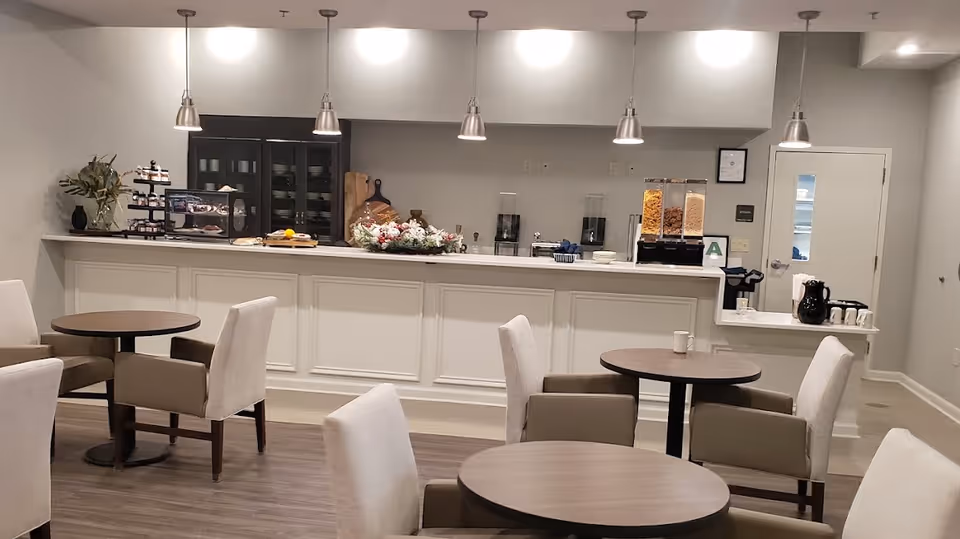 A clean and modern dining area with round tables and cushioned chairs. In the background, there is a counter with various food items, cereal dispensers, and coffee pots. Pendant lights hang from the ceiling illuminating the space.