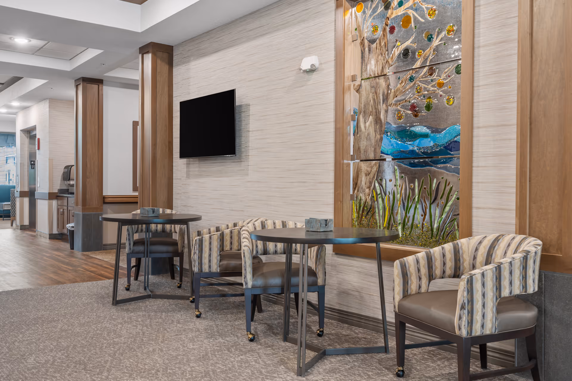 A seating area in a senior living facility with two round tables and four cushioned chairs with patterned upholstery. The wall behind features a mounted flat-screen TV and a decorative glass artwork depicting a tree with colorful leaves and a blue river. The floor is carpeted near the seating area and transitions to wood flooring in the hallway.