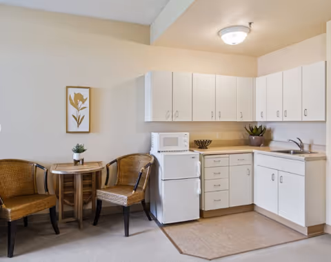 Small kitchenette with white cabinets, a mini-fridge and microwave, sink, and a small table with two wicker chairs.