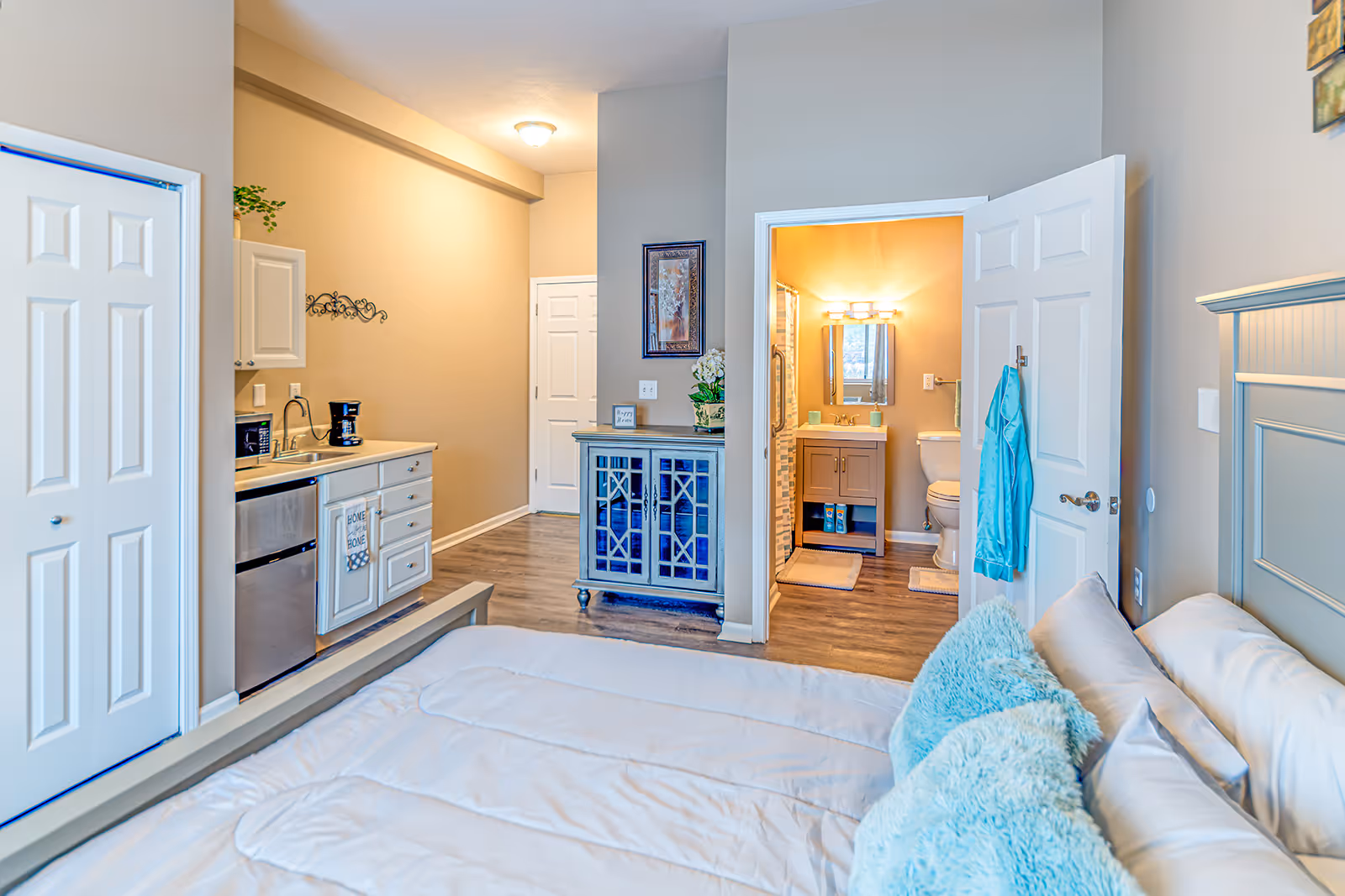 Bright studio apartment bedroom with a bed in the foreground, a small kitchenette to the left, and an open bathroom door straight ahead.