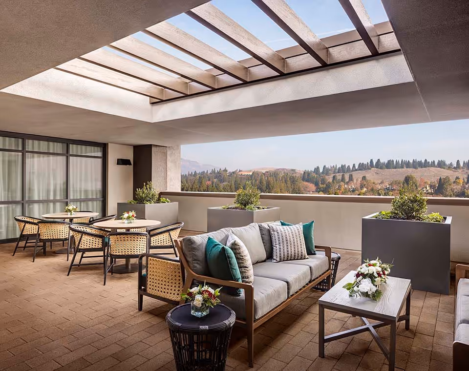 Outdoor covered patio area with comfortable seating including a sofa with cushions, a coffee table with floral arrangements, and round tables with chairs. Large planters with greenery line the edge of the patio, overlooking a scenic view of hills and trees under a partly cloudy sky.