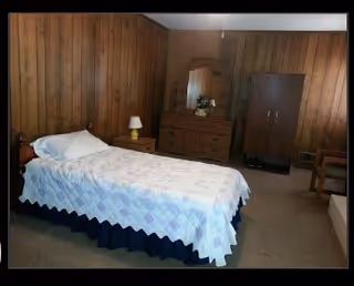 A simple bedroom with a single bed covered by a quilt, wood-paneled walls, a dresser with mirror, wardrobe, nightstand and lamp.