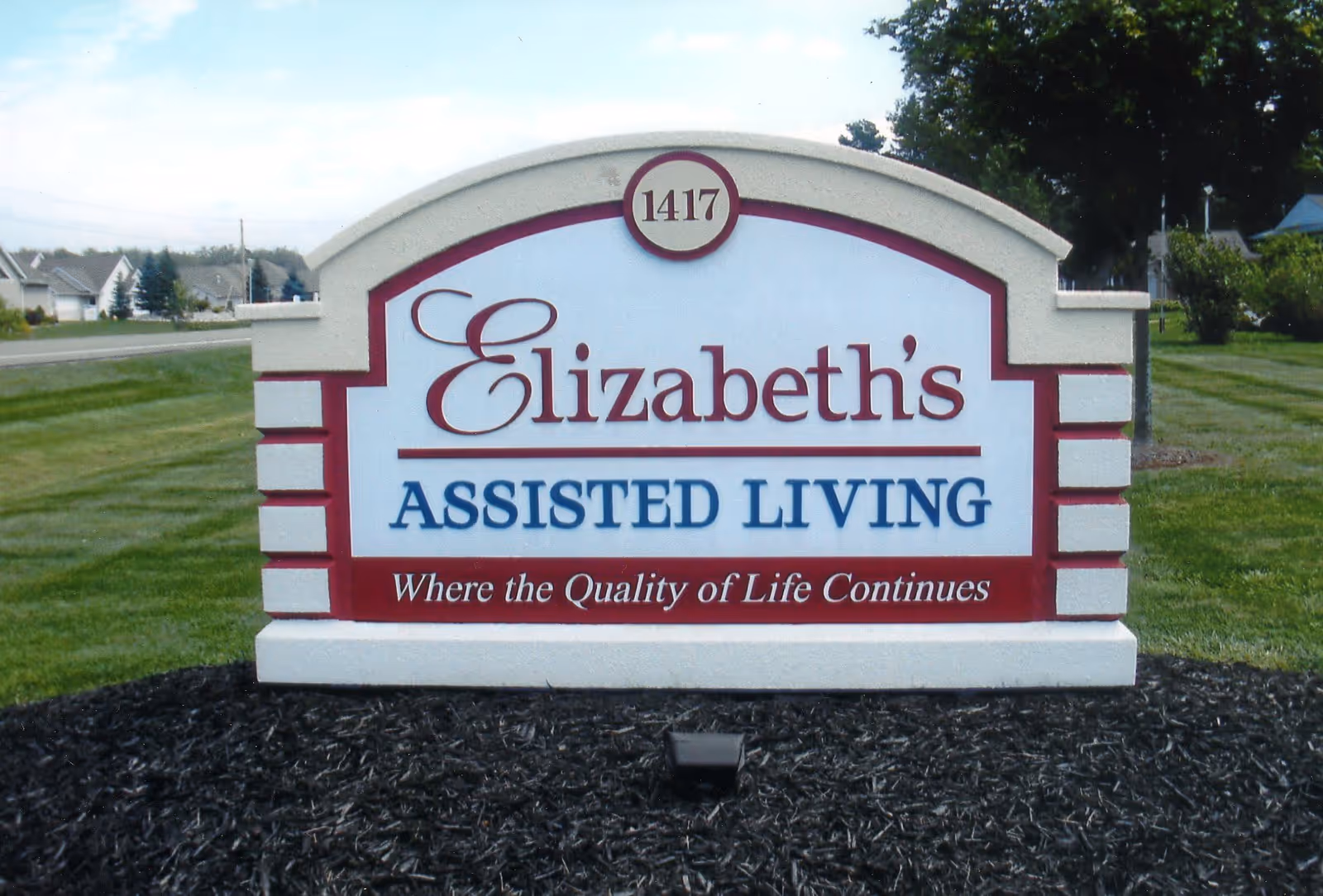 A large outdoor sign for Elizabeth's Assisted Living with the address number 1417 displayed at the top. The sign includes the tagline 'Where the Quality of Life Continues' and is set on a landscaped area with grass and mulch.