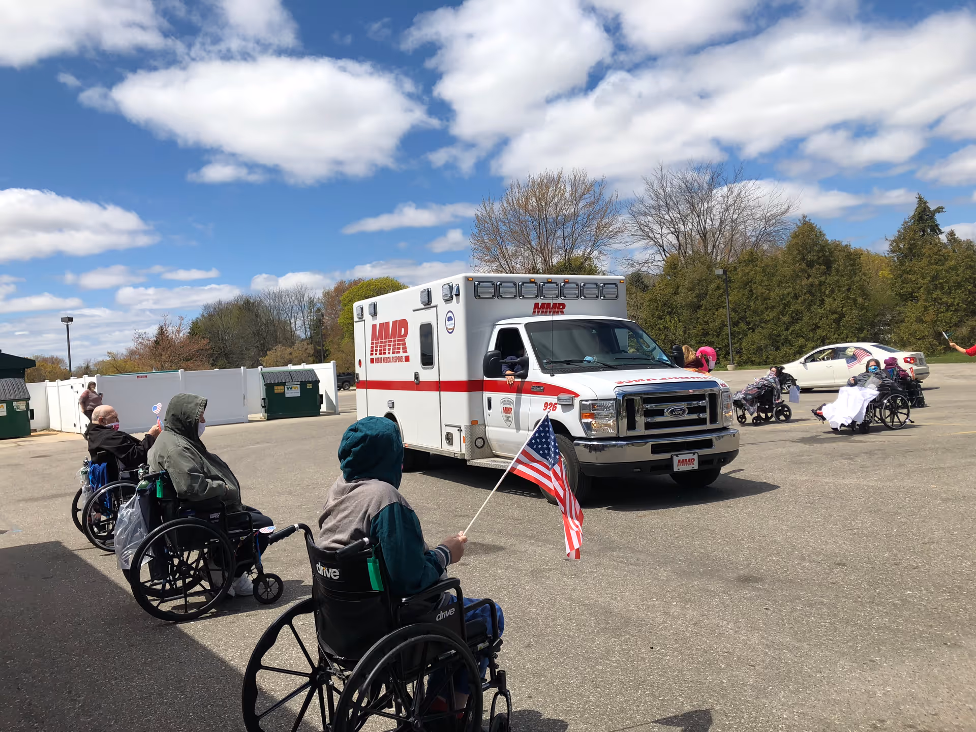 Several elderly individuals in wheelchairs are outside in a parking lot, some holding American flags, watching a white ambulance with red stripes and MMR written on it. The sky is partly cloudy and there are trees and a white fence in the background.