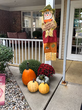 Front porch area decorated for fall with a scarecrow holding a 'Happy Harvest' sign, surrounded by pumpkins, gourds, and potted chrysanthemums. There is a white railing, a seating area with cushions, and a glass door leading inside the building.