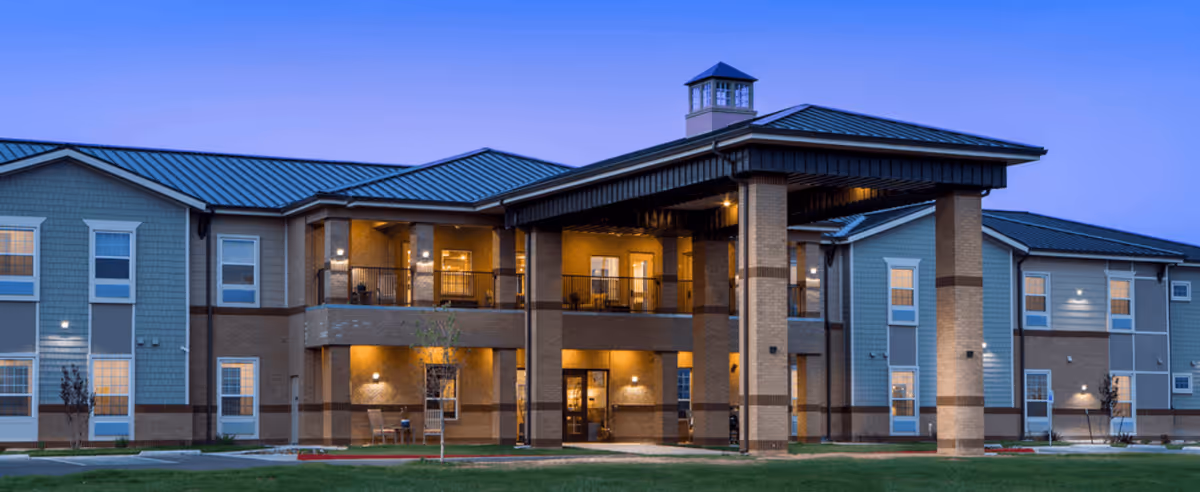Exterior view of The Legacy at South Plains senior living facility at dusk, showing a two-story building with lit windows and a covered entrance supported by brick columns.