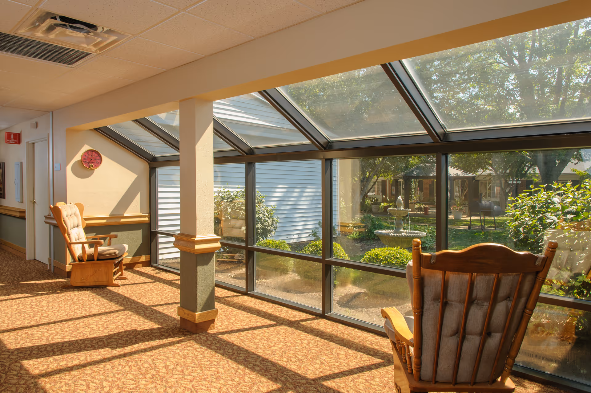 Sunlit indoor seating area with wooden rocking chairs along a glass-enclosed corridor overlooking a courtyard fountain.