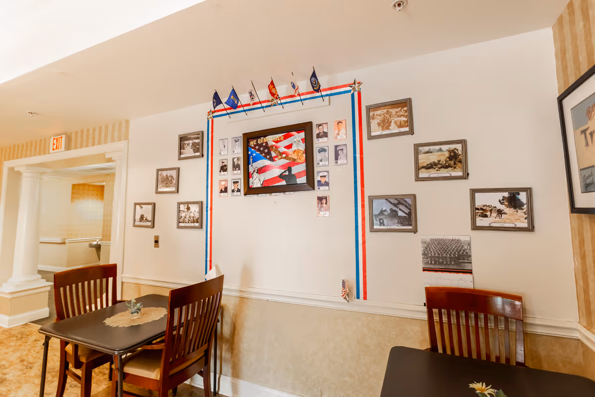 Small communal dining area with tables and wooden chairs facing a wall decorated with framed photos, flags, and patriotic ribbons.