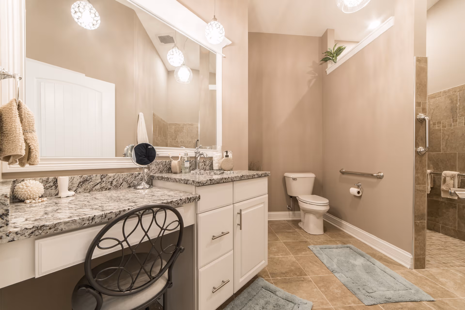 A spacious bathroom with beige walls and tiled floor featuring a white toilet, a walk-in tiled shower with grab bars, a granite countertop vanity with a large mirror, and a black metal chair with a cushion. Two blue bath mats are placed on the floor, and there are decorative items and towels on the countertop and hanging on the wall.