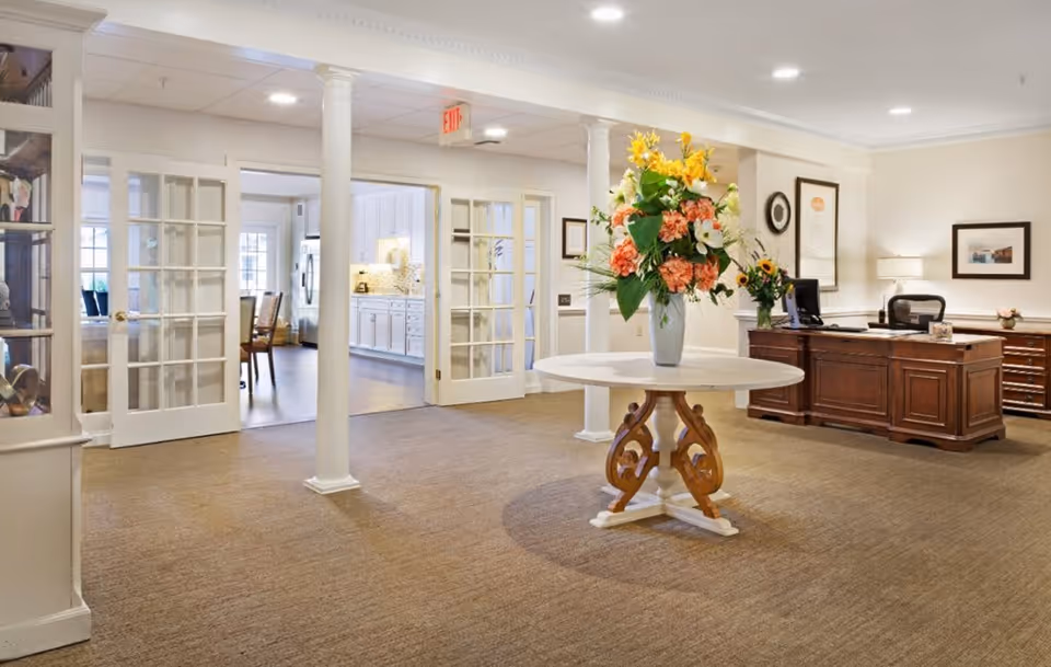 A bright and welcoming interior space featuring a round table with a large floral arrangement in the center. To the right, there is a wooden reception desk with a computer and lamp. The background shows glass-paneled double doors leading to a kitchen area with white cabinetry and dining chairs. The room has beige walls, carpeted flooring, and white columns supporting the ceiling.