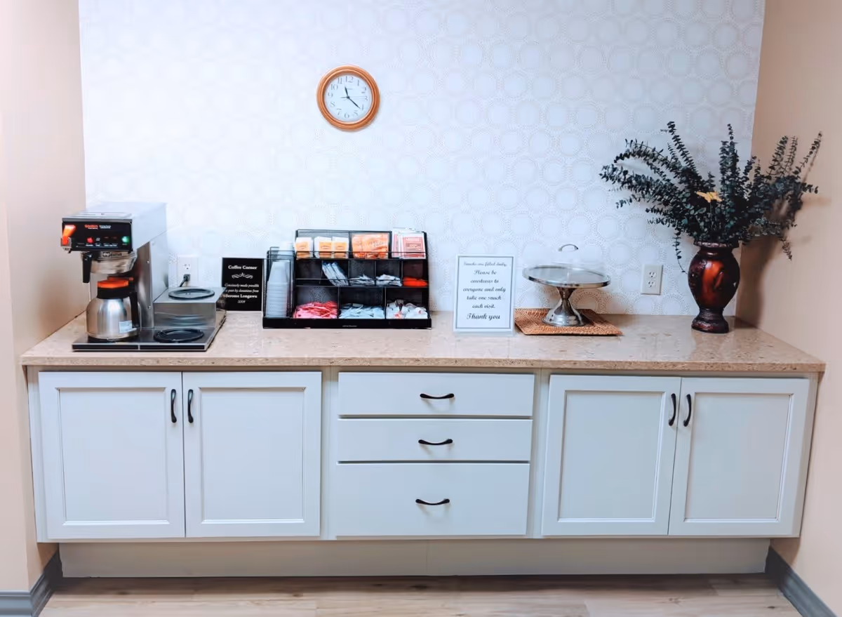 A coffee station in a room with a beige countertop and white cabinets below. On the left side, there is a coffee maker with a pot of coffee. In the center, there is a black organizer holding coffee supplies such as cups, sugar packets, and stirrers. A small sign is placed next to the organizer. On the right side, there is a covered cake stand and a vase with green foliage. A round wall clock with a wooden frame hangs on the patterned white wall above the countertop.