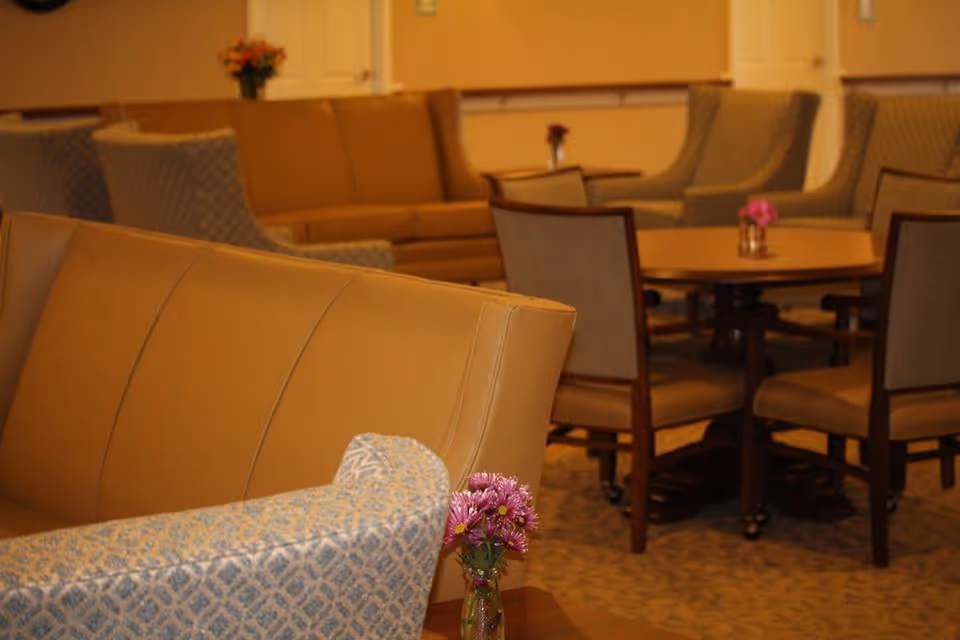 Interior view of a senior living facility lounge area with tan leather sofas, patterned armchairs, and a round wooden table surrounded by chairs. Small vases with flowers are placed on the tables.