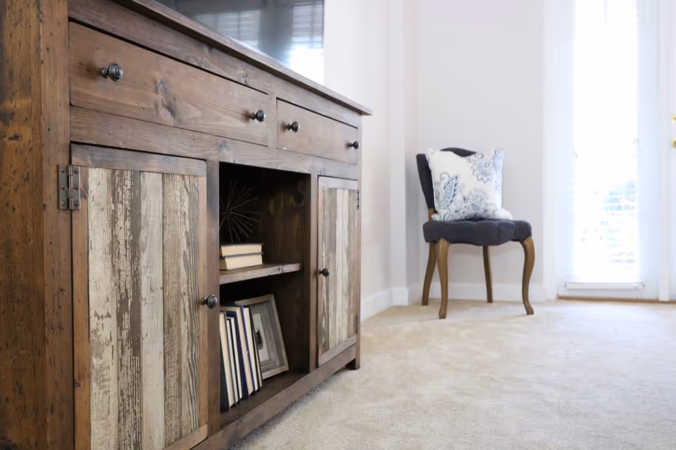 A rustic wooden cabinet with drawers and shelves holding books and a picture frame, next to a dark upholstered chair with a decorative pillow, set on a carpeted floor near a window with blinds.