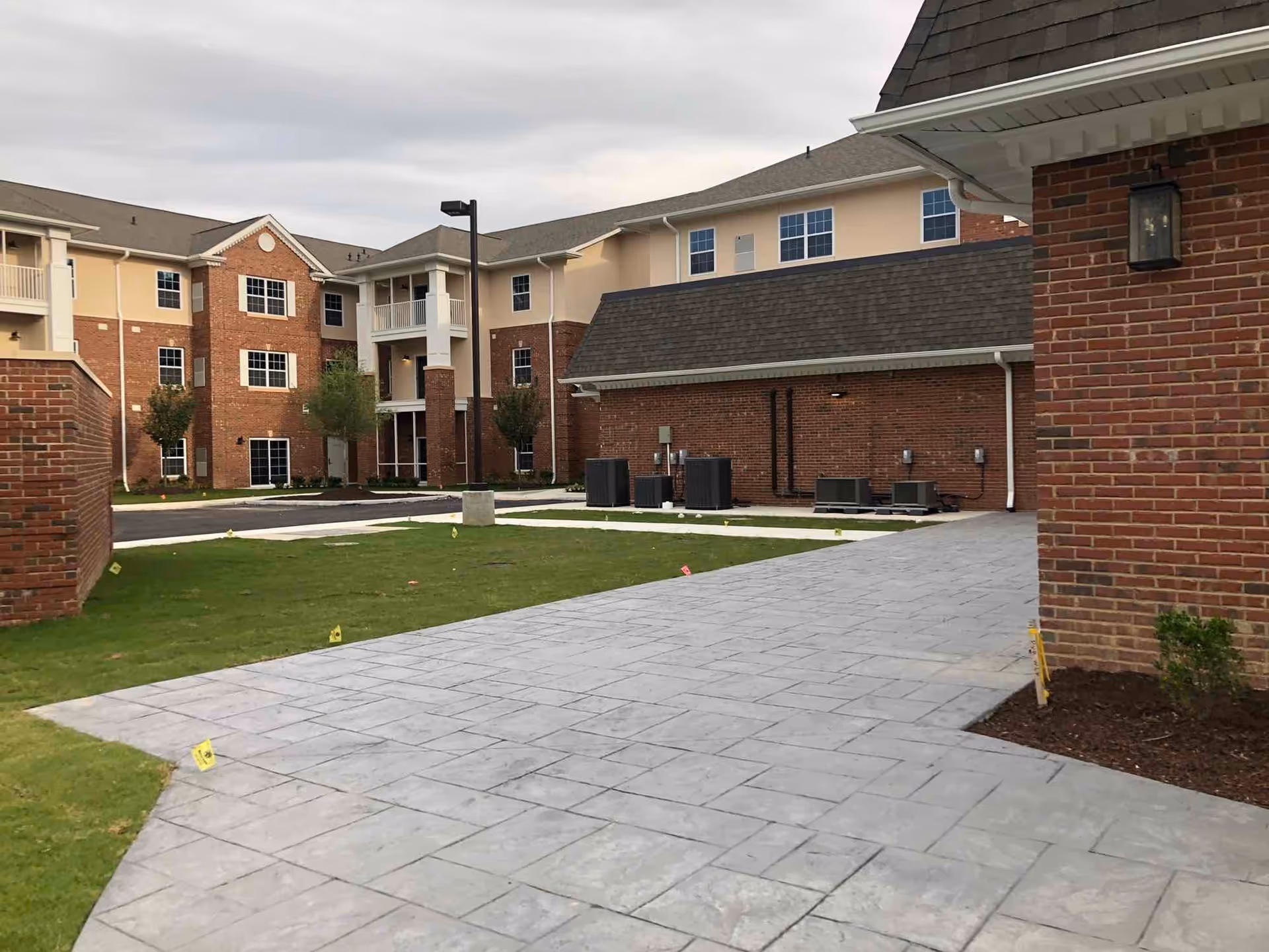 Exterior view of a brick multi-story residential building with a paved walkway, small lawn, and outdoor HVAC units.