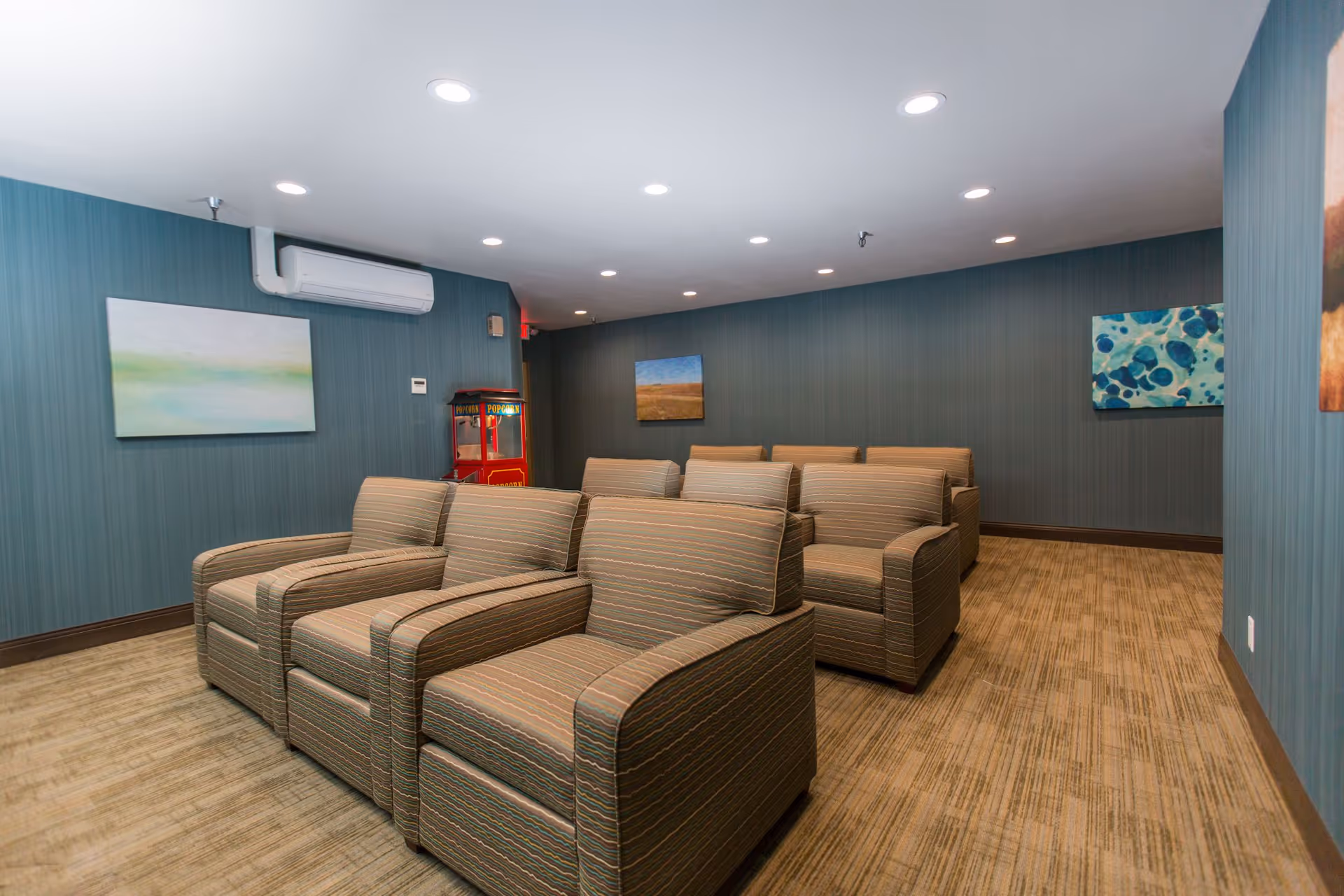 A small theater room with two rows of comfortable striped armchairs facing forward. The walls are painted blue with several abstract paintings hanging. There is a red popcorn machine in the back corner and recessed ceiling lights illuminating the room.