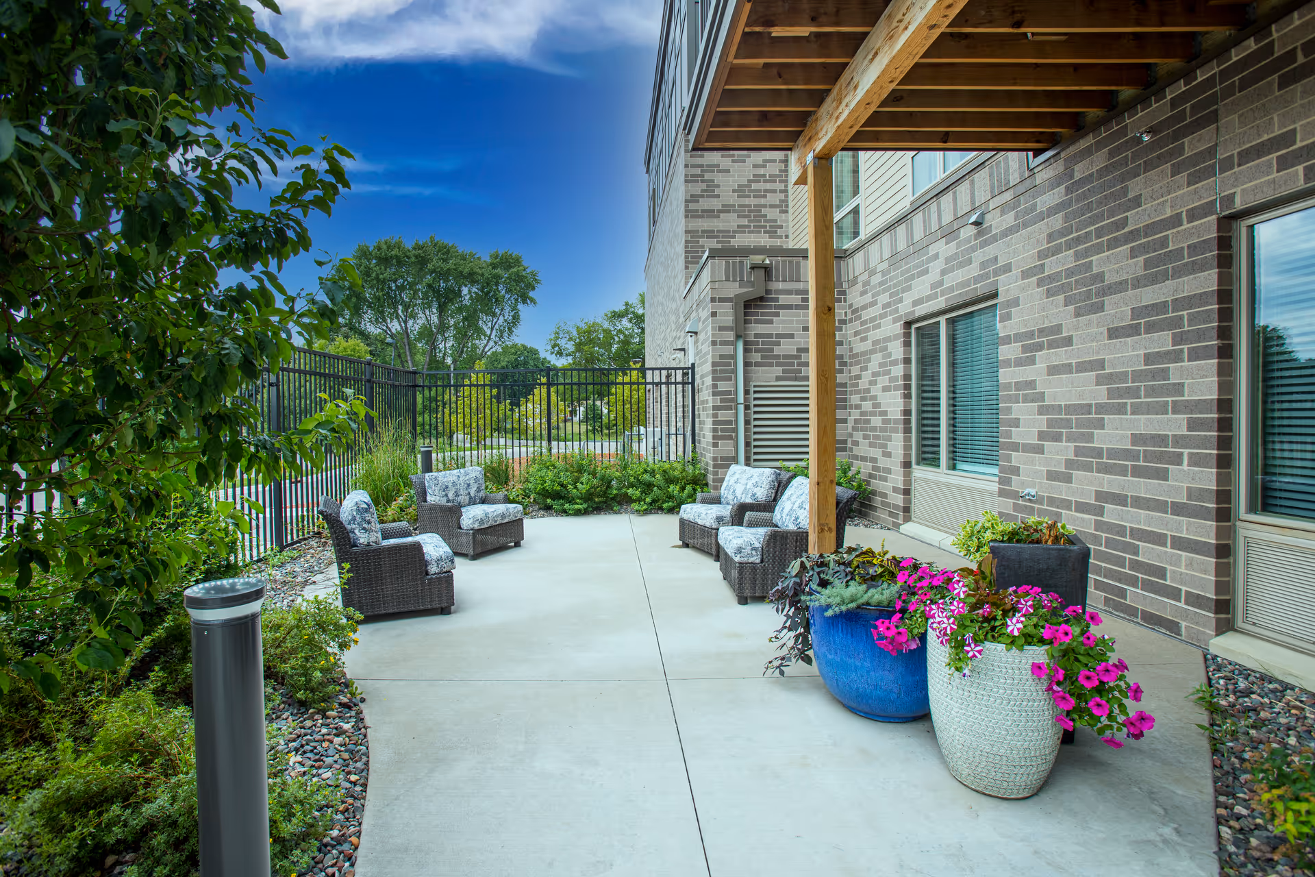 Outdoor patio area at a senior living facility with cushioned wicker chairs arranged on a concrete surface, surrounded by potted plants and greenery, next to a brick building under a wooden pergola.