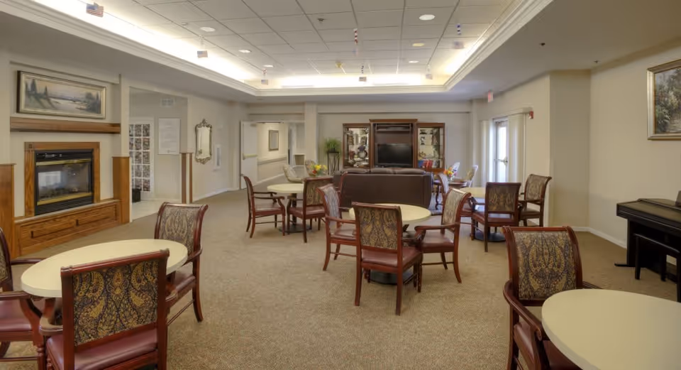 A spacious senior living common area with multiple round tables and patterned chairs arranged throughout the room. There is a fireplace with a wooden mantle on the left wall, a large entertainment center with a TV in the back center, and a piano on the right side. The room has beige walls, carpeted floors, and a drop ceiling with recessed lighting.