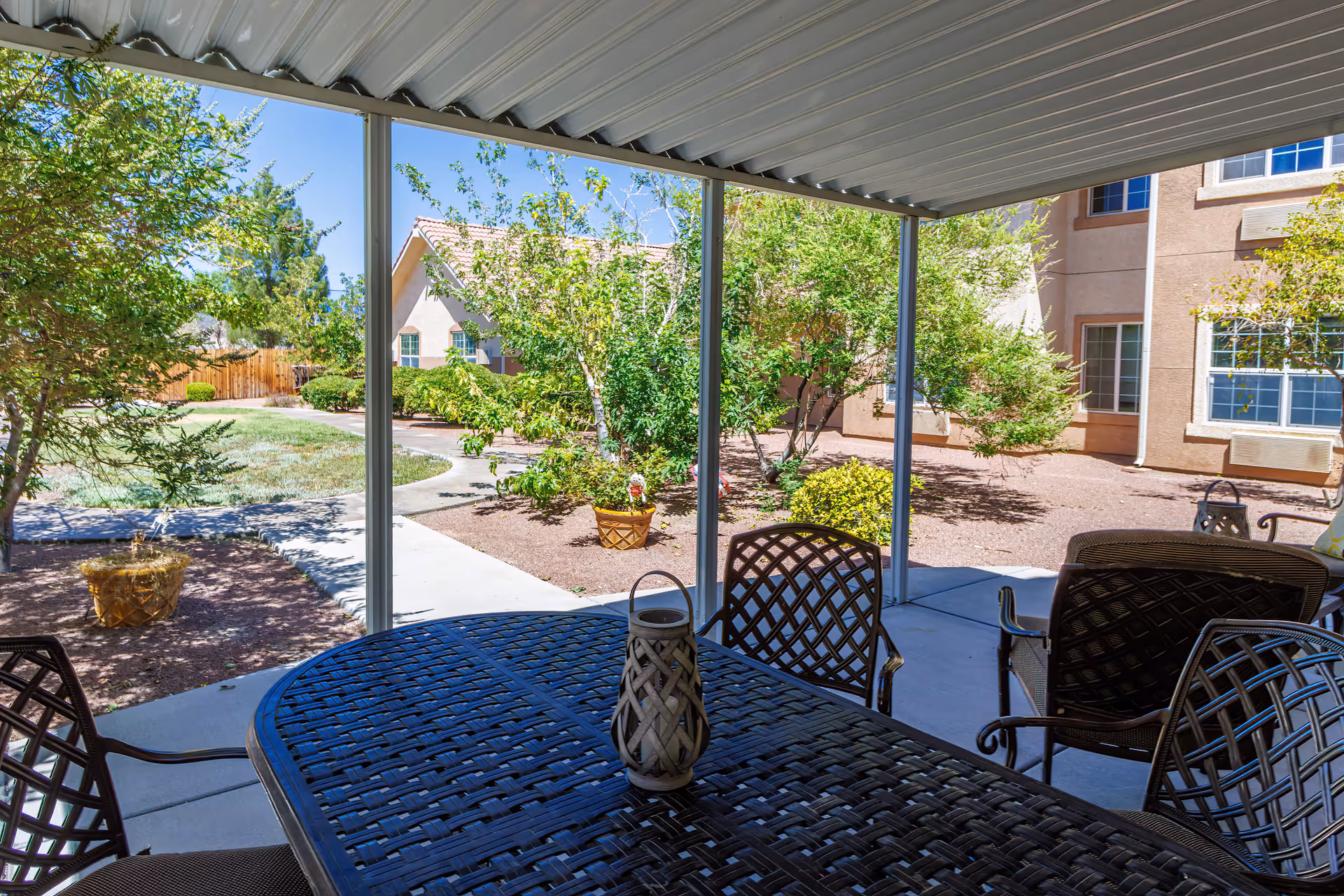 Covered outdoor patio area with a metal table and chairs. The patio overlooks a landscaped garden with trees, shrubs, and a walking path. In the background, there are beige buildings with windows under a clear blue sky.