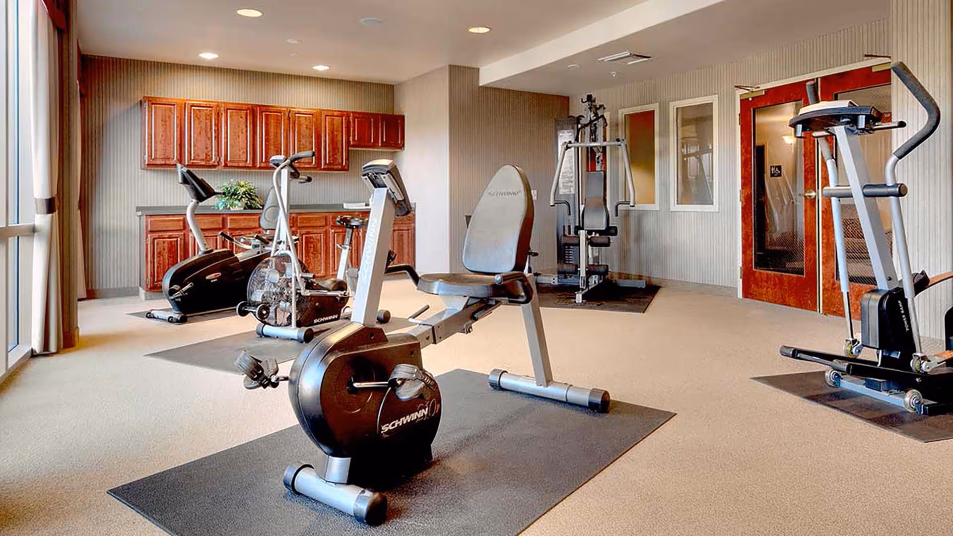 A fitness room with exercise equipment including stationary bikes, a multi-function weight machine, and an elliptical trainer. The room has beige carpet, light-colored walls with vertical stripes, wooden cabinets along one wall, and large windows with curtains letting in natural light.