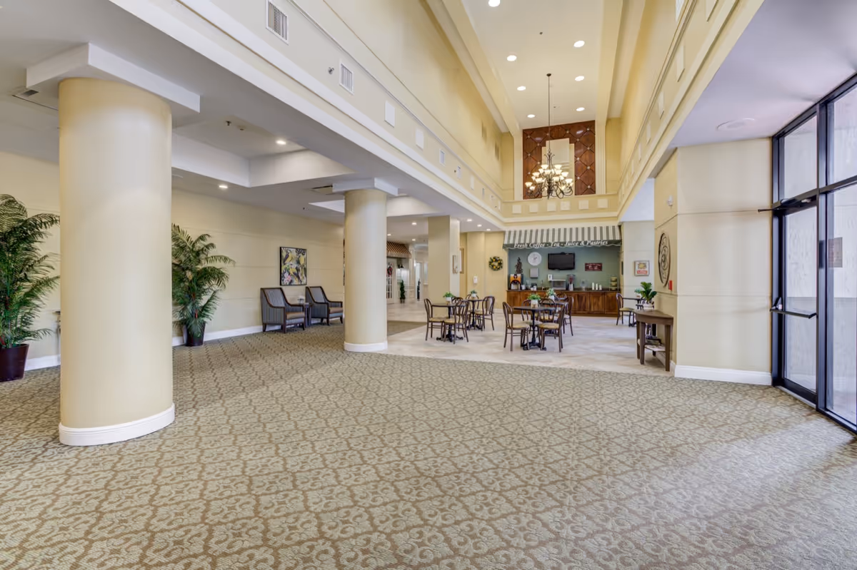 Spacious lobby of a senior living facility with columns, seating areas, and a café-style dining area by large glass doors.