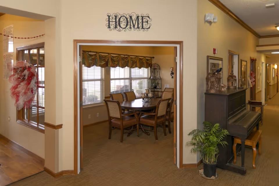 Hallway opening into a dining room with a round wooden table and chairs, a piano and potted plant along the corridor.