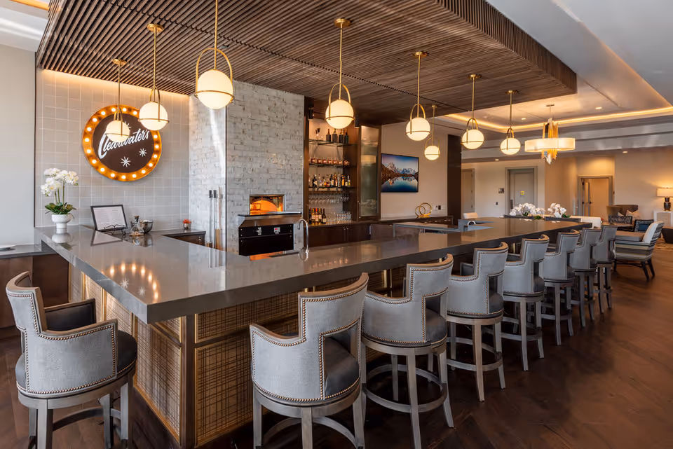 Modern communal bar and lounge with a long stone countertop, a row of upholstered bar stools, pendant lights, and wood accents.