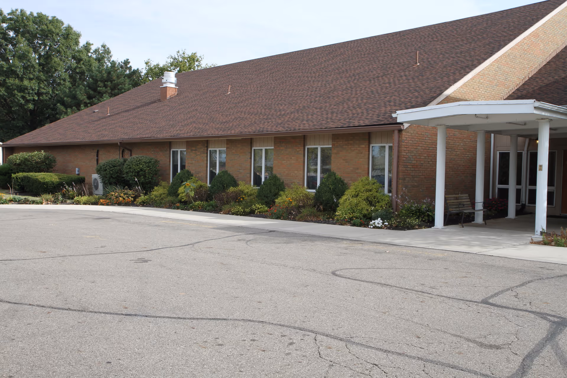 Single-story brick building with a covered entrance, shrubs and flowerbeds along the facade and a paved driveway.