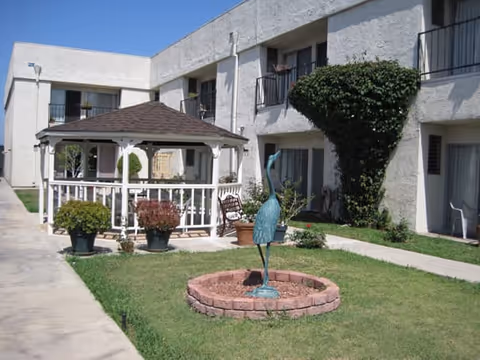 Outdoor courtyard area of a retirement center featuring a white gazebo with a brown roof, potted plants, a metal bird sculpture in a circular brick planter, and a two-story building with balconies and windows in the background under a clear blue sky.