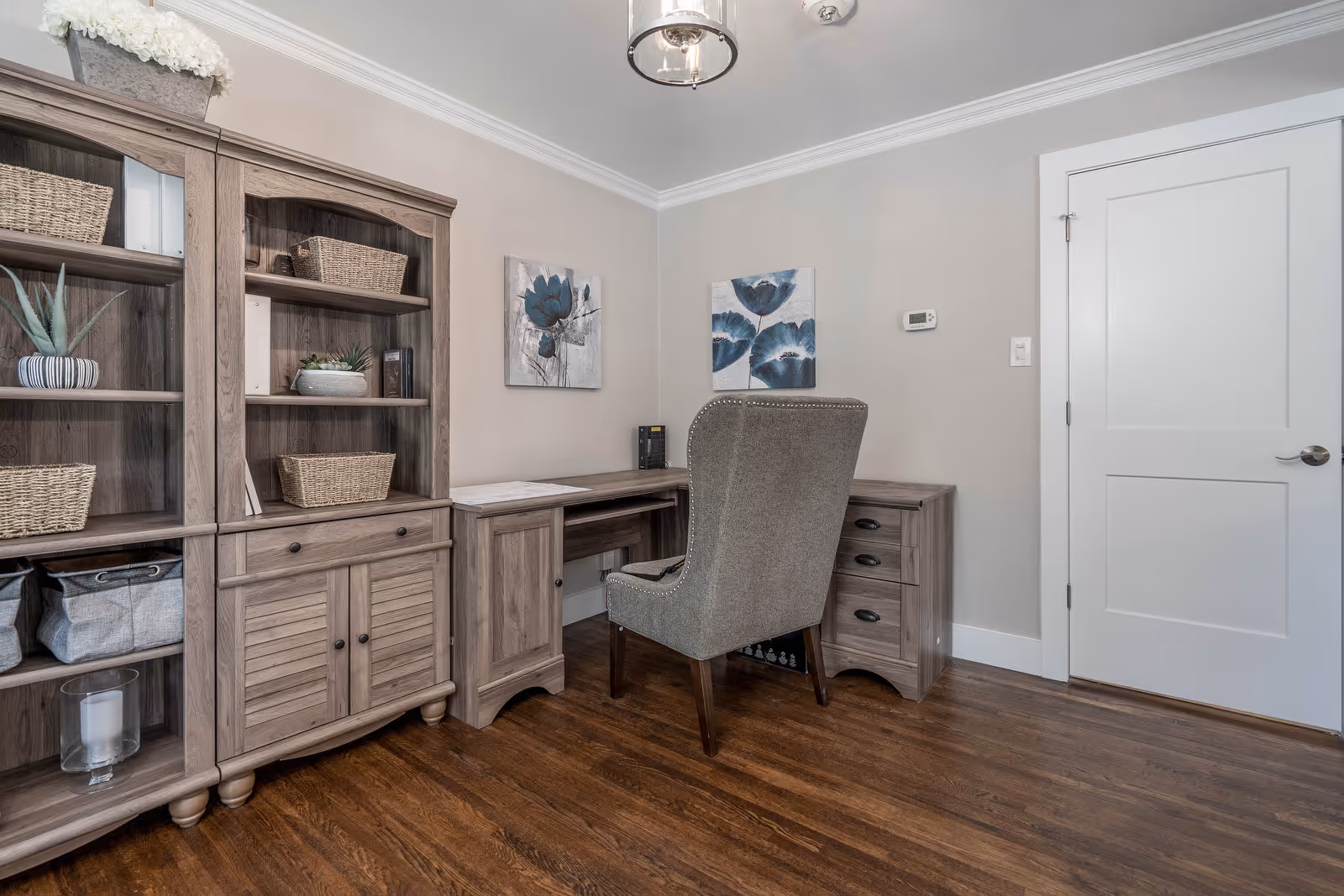 A home office with a wooden desk and matching wooden shelving unit filled with baskets and decorative items. A gray upholstered chair is positioned at the desk. Two floral paintings hang on the beige wall above the desk. The room has hardwood flooring and a white door with a silver handle.