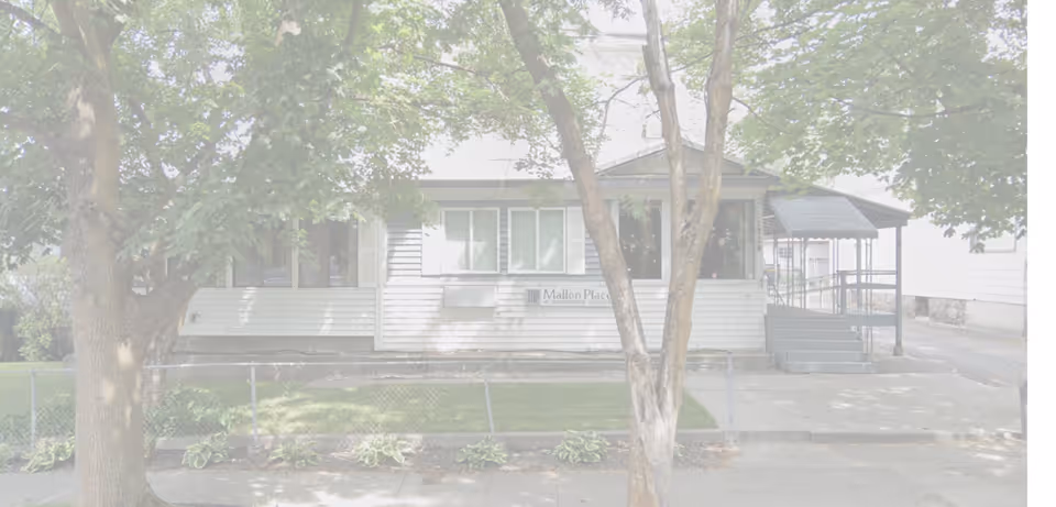 Front exterior of Mallon Place, a single-story house-like building with a covered ramp entrance, trees and a chain-link fence.