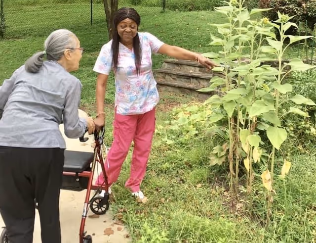 An elderly woman using a walker is accompanied by a caregiver wearing pink scrubs. They are outside near a garden area with green plants and a small set of wooden steps in the background.