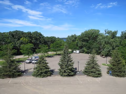 View over a mostly empty parking lot with several evergreen trees, a few parked vehicles, and a wooded area with a distant lake under a blue sky.