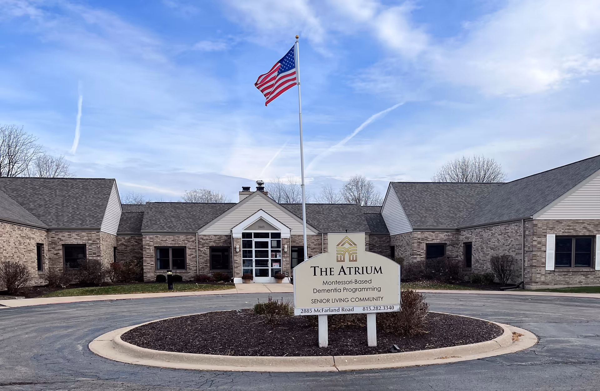 Front exterior of The Atrium senior living community with a sign, flagpole, and circular driveway.