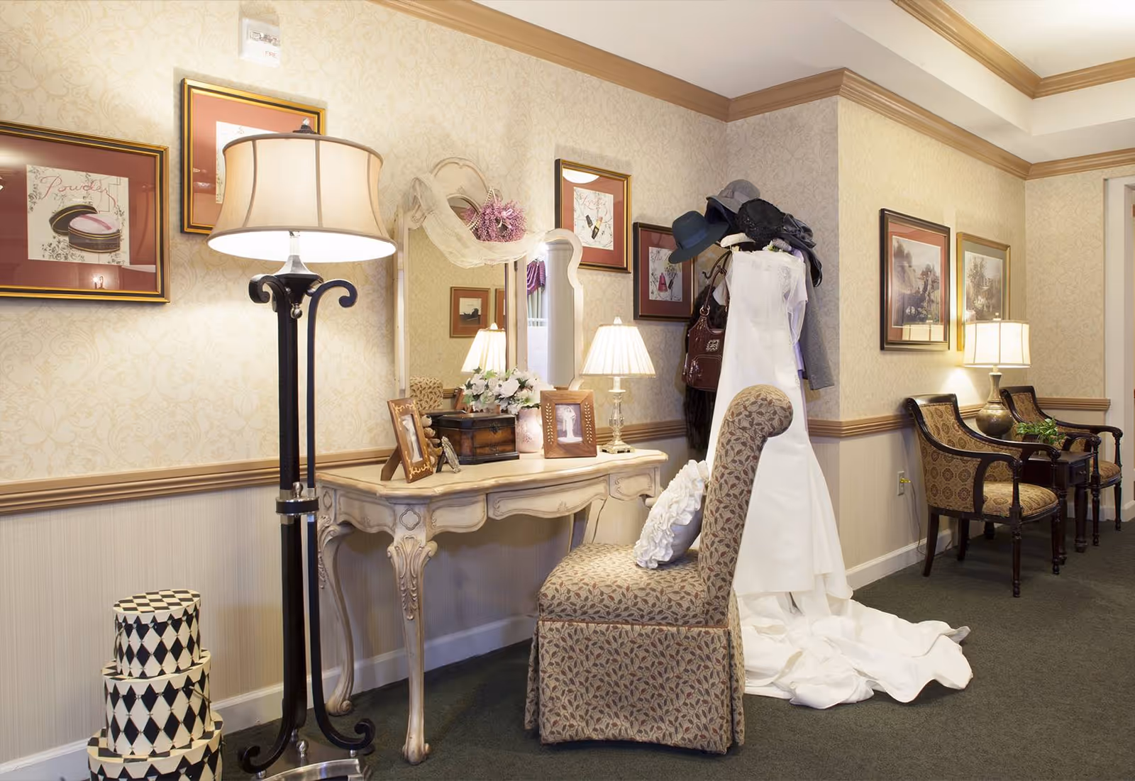 A cozy interior corner of a senior living facility with a vintage-style vanity table and mirror, a cushioned chair, a floor lamp, and a mannequin dressed in a white gown with hats and accessories. The walls are decorated with framed artwork and wallpaper, and there are additional chairs and a small table with a lamp in the background.
