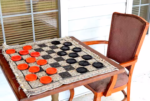 A wooden table with a woven checkerboard and red and black checkers set up for a game, next to a brown cushioned wooden chair on a porch with white siding and a window with blinds.
