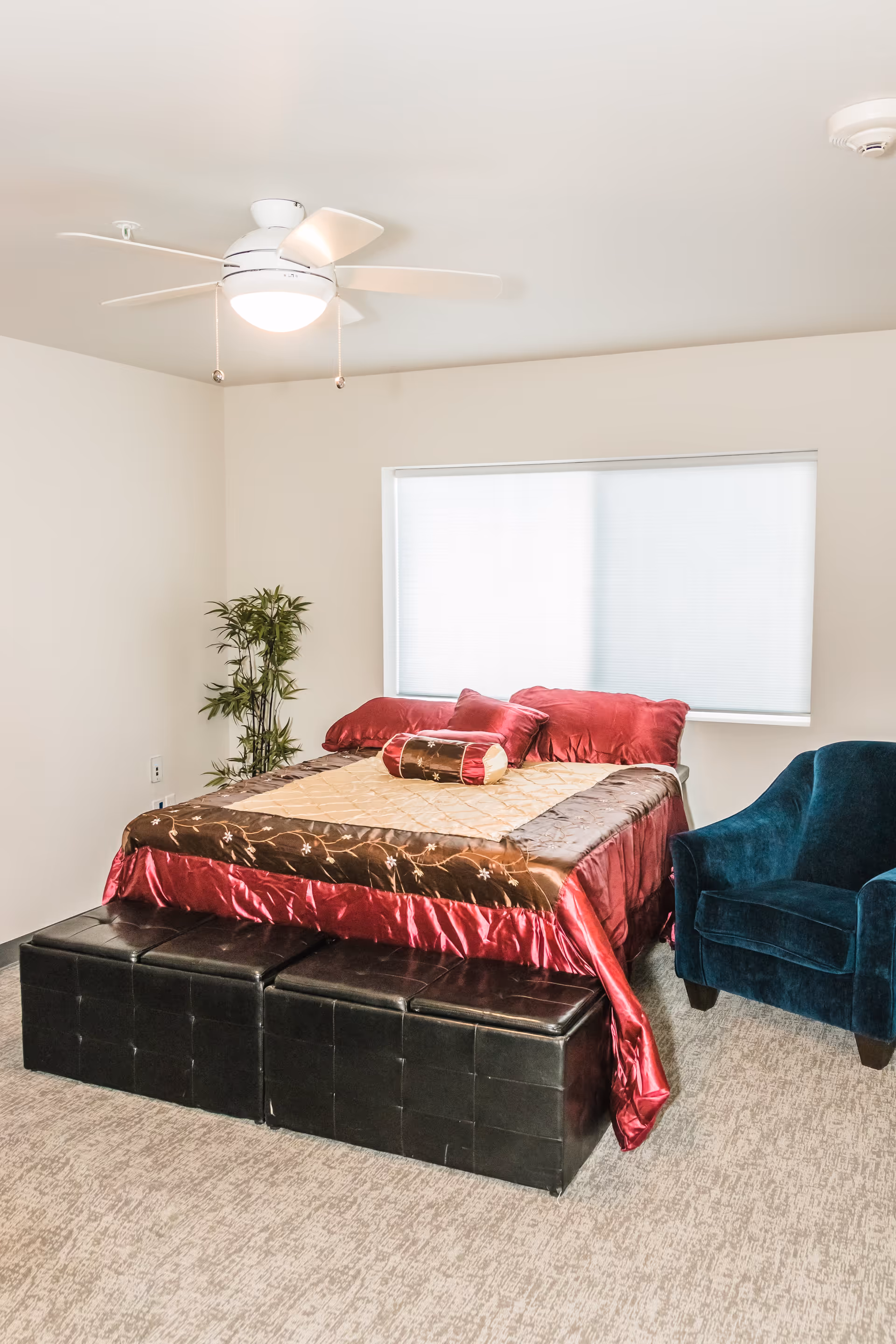 A bedroom with a bed covered in a red and brown quilt with matching pillows. At the foot of the bed are two black ottomans. To the right of the bed is a dark blue upholstered armchair. A ceiling fan with a light is mounted on the white ceiling, and a window with a white blind is on the wall behind the bed. A small green plant is in the corner of the room.
