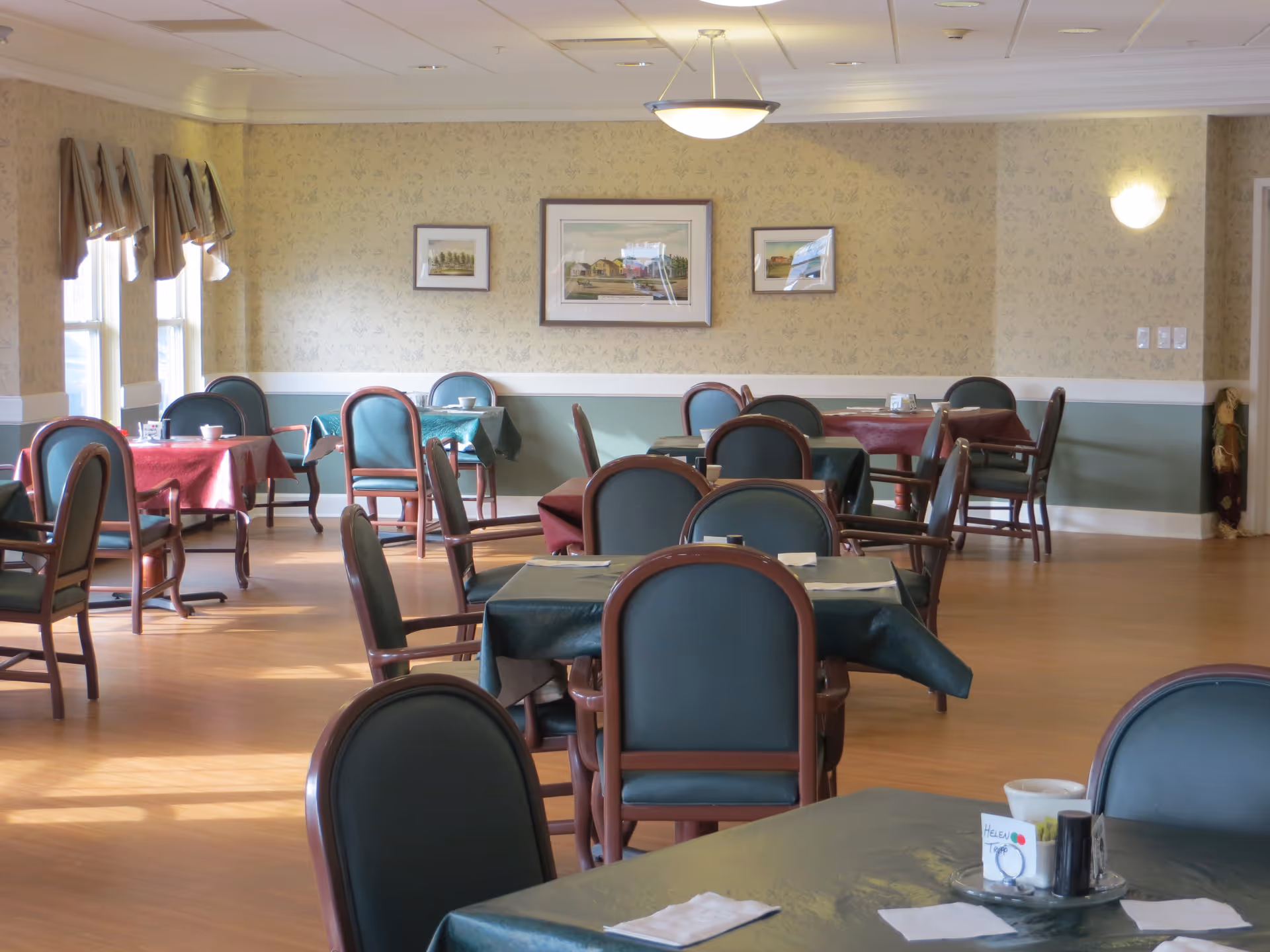 Empty senior living dining room with tables covered in tablecloths and green upholstered chairs arranged around the room.