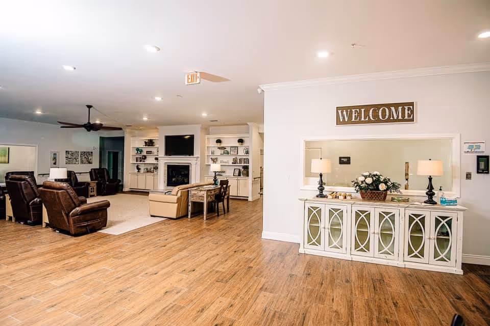 Bright communal living area with leather recliners, a fireplace and TV, shelving, and a 'WELCOME' sign over a console table.