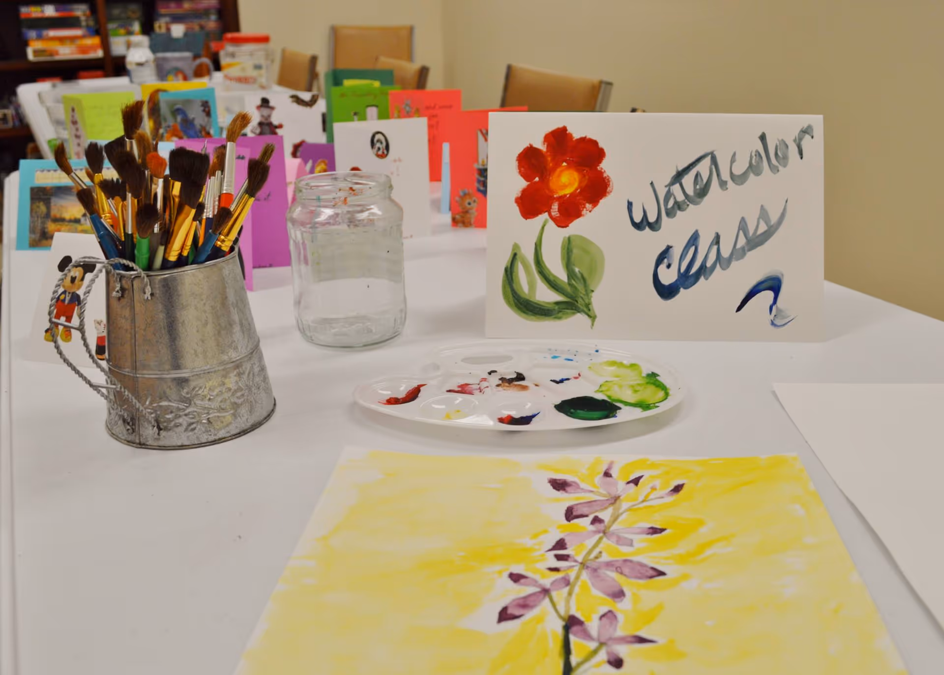Table set up for a watercolor class with brushes in a metal container, a paint palette and jar, and a card labeled "watercolor class".