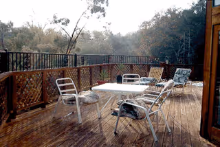 Wooden outdoor deck with a white table and several metal chairs with cushions. The deck is surrounded by a wooden lattice railing and overlooks a wooded area with trees.