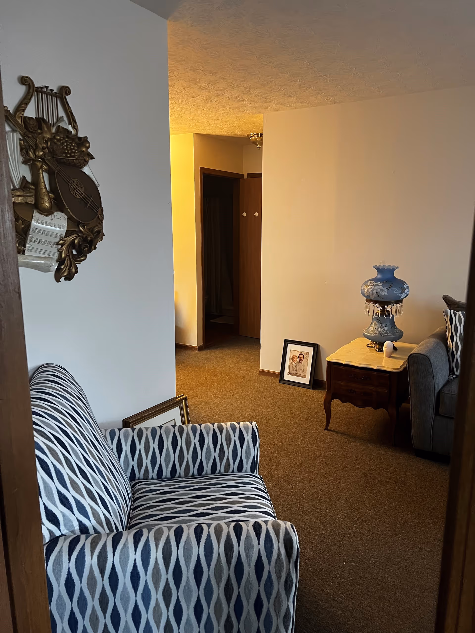 Interior view of a living room area with a patterned armchair in the foreground, a wooden side table with a decorative blue lamp, and a gray sofa partially visible. There are framed pictures resting on the carpeted floor near the wall and an open doorway leading to another room.